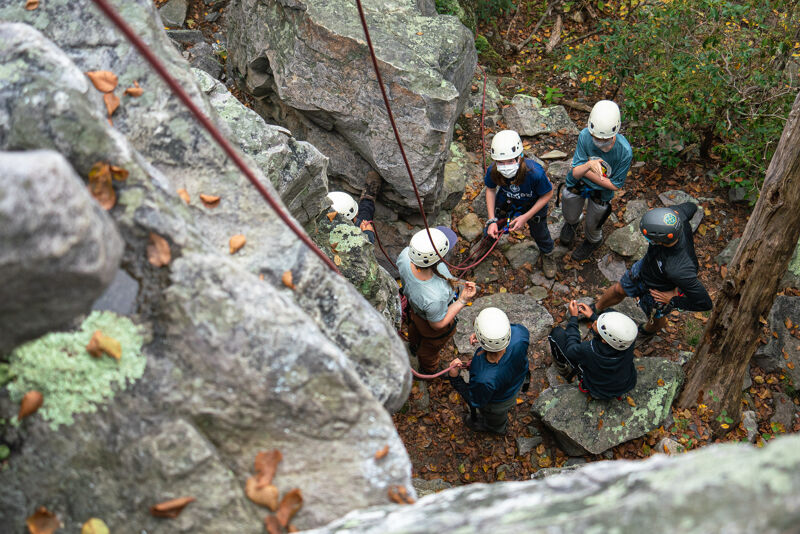 A group of people wearing helmets are gathered around a rocky area, possibly preparing for or engaging in rock climbing or rappelling. Ropes are visible, suggesting climbing equipment. The setting appears to be outdoors, with trees and foliage in the background, indicating a natural environment.
